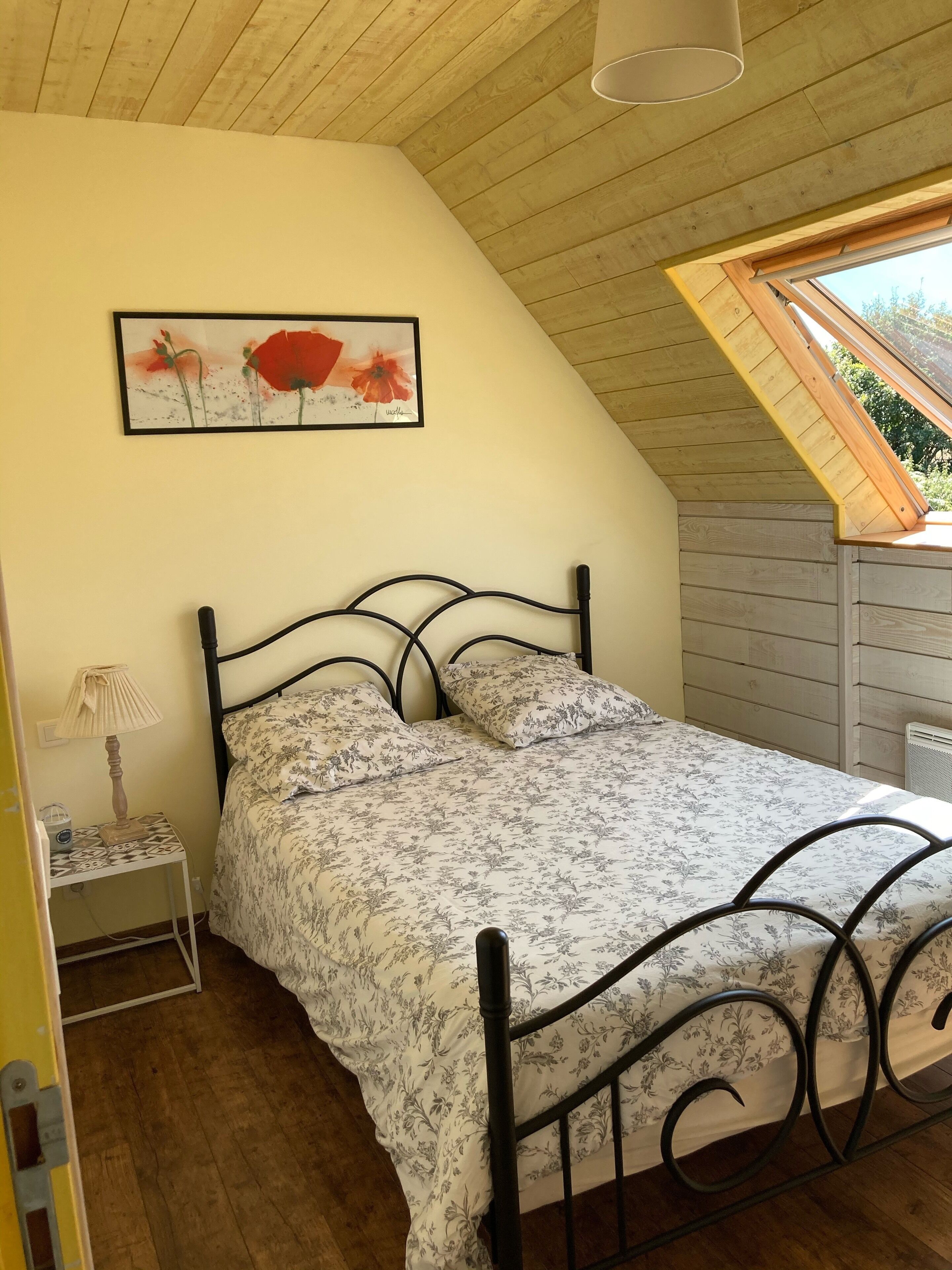 Une chambre avec un plafond en pente, un lit recouvert d’une couvre-lit à motifs floraux, une table de chevet avec une lampe et un tableau encadré accroché au mur.