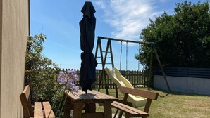 A wooden deck with picnic tables and chairs, a swing set, and a covered umbrella. - Gîte de Penn Ar Roz: 3 épis seaside GUILER-SUR-GOYEN, 4-5 pers, swimming pool. (Guiler-sur-Goyen)
