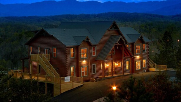 A large log cabin with multiple windows and a staircase, surrounded by a forested landscape.
