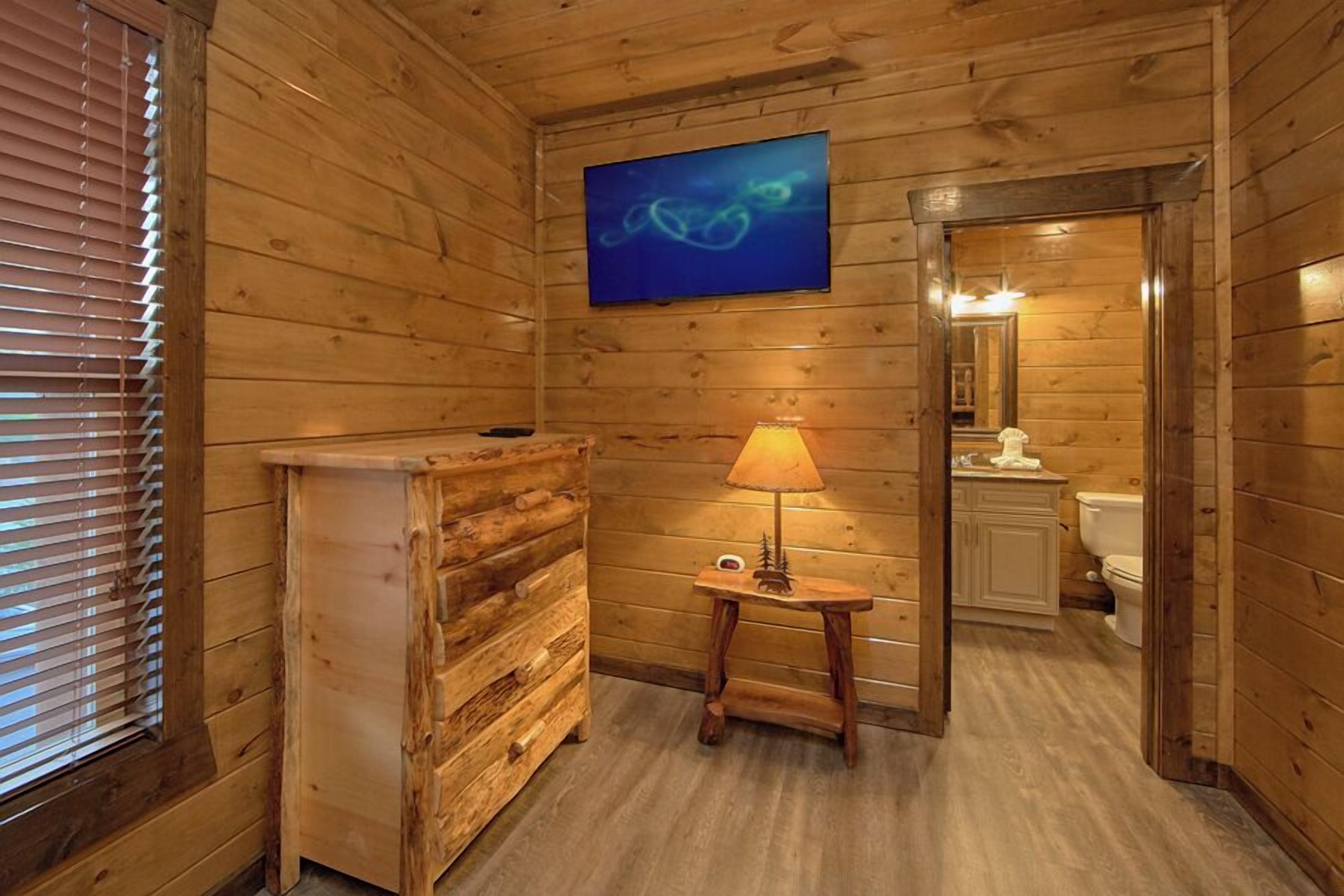 Un baño de cabaña de madera con un banco de madera, una mesita con lámpara, un televisor montado en la pared y un espejo en la habitación contigua.