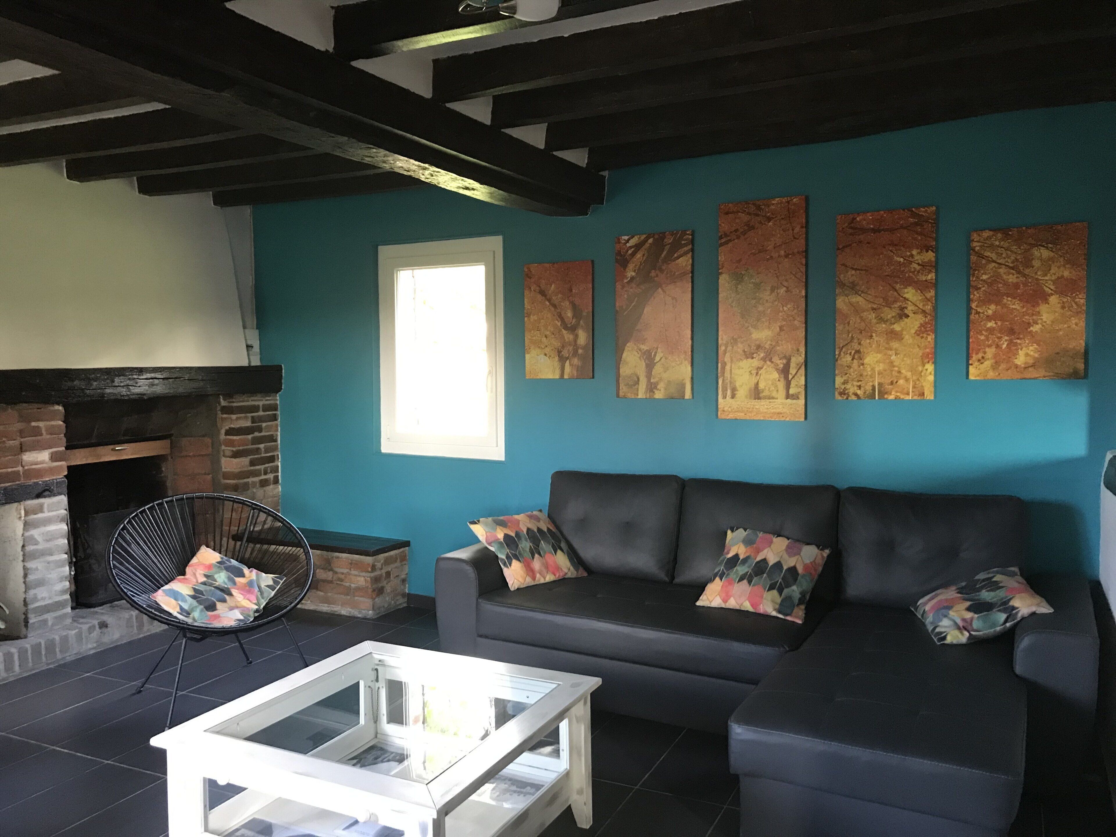 A living room with a black sofa, colorful cushions, a glass coffee table, a black chair with a colorful cushion, a fireplace, and four autumn-themed paintings on the wall.