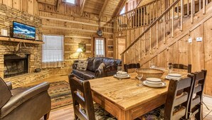 Ein rustikales Holzinterieur mit Kamin aus Stein, Ledersofa, Esstisch und einer Holztreppe.