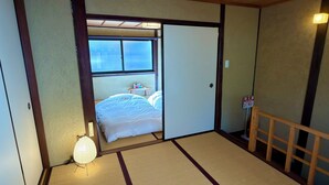 Una habitación japonesa tradicional con suelo de tatami, una cama con ropa blanca, una mesita con objetos y un puertas correderas que dan a un balcón.