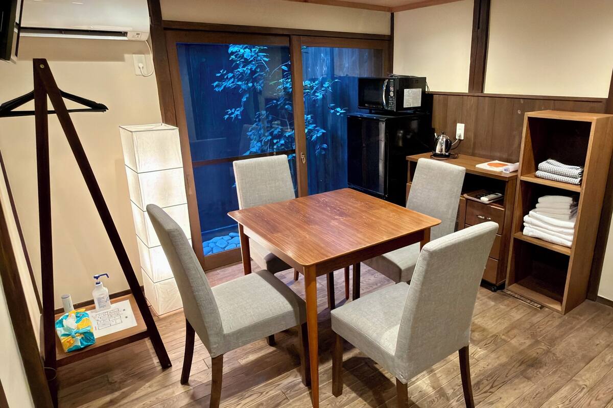 A compact room with a wooden table and four chairs, a microwave, a small refrigerator, a wooden wardrobe, and a sliding glass door with a view of blue foliage.