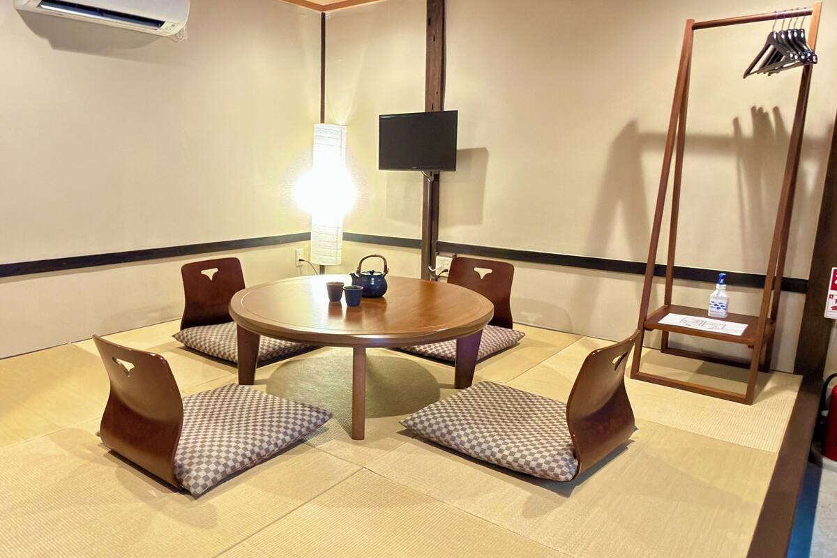 A conference room with a round table, four chairs, a flat-screen TV, a standing lamp, and a small shelf with a bottle.
