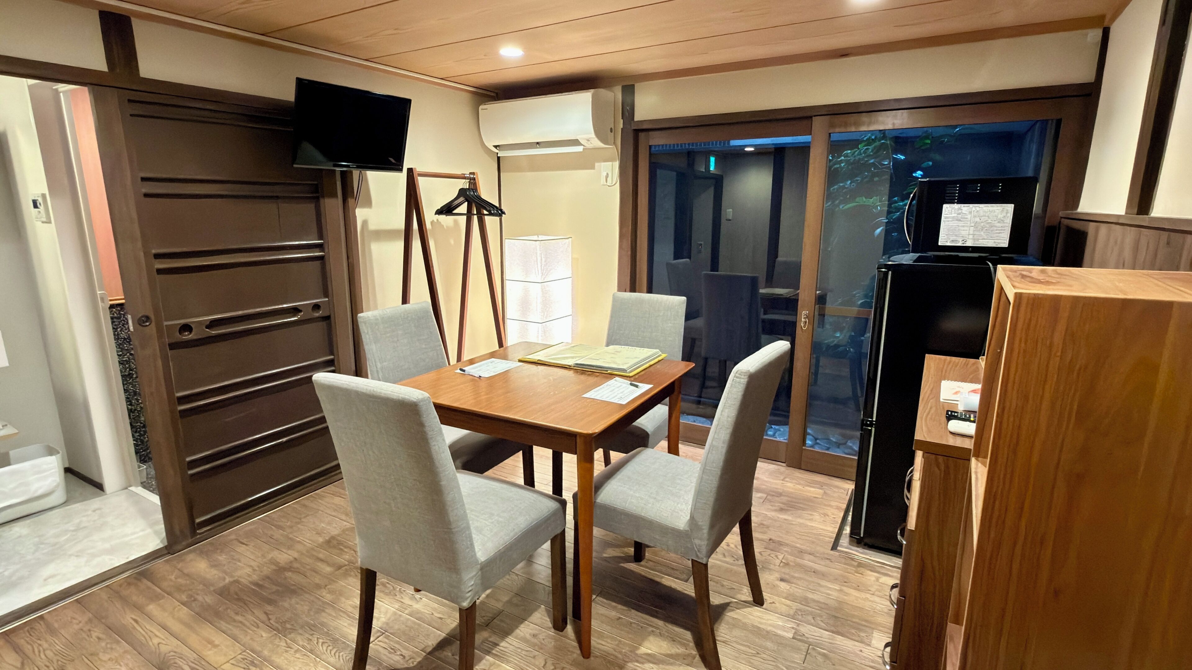 A dining area with a wooden table, grey upholstered chairs, a television mounted on a wall, and a refrigerator.