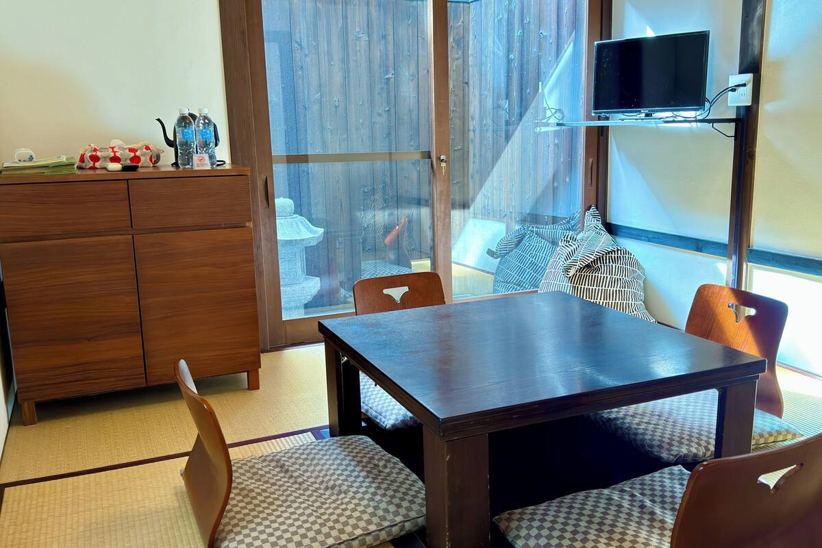 A room with a wooden table, chairs, a TV on a stand, a cabinet, and a view of a garden through a glass door.