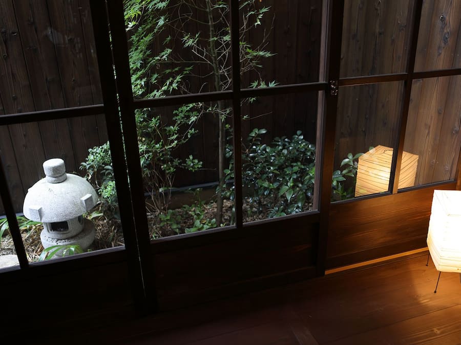 A room with a view through a wooden-framed window, a stone lantern, and a stack of papers on the floor.