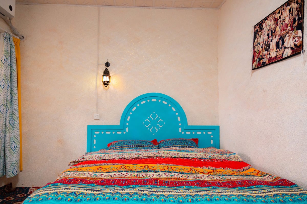 Quarto com cama, cabeceira azul, roupa de cama estampada, luminária fixada na parede e quadro na parede.