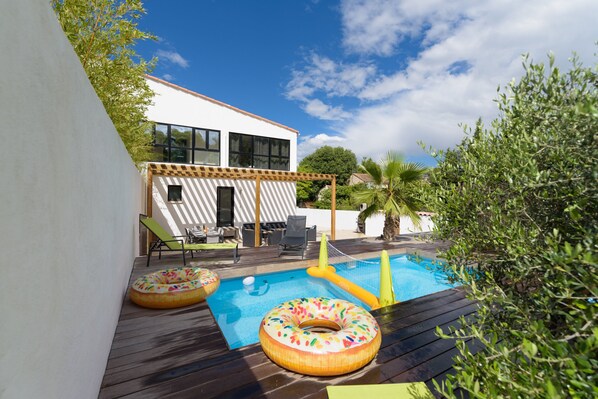 Una piscina exterior con flotadores inflables, una terraza de madera y un edificio blanco con amplias ventanas.