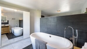 Un baño moderno con una bañera independiente, un espejo grande y paredes cubiertas de baldosas oscuras.
