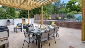 Un patio exterior con pérgola, mesa de comedor preparada para cuatro personas y una parrilla de barbacoa al fondo.