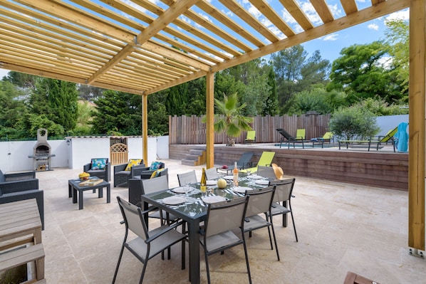 Un patio exterior con pérgola, mesa de comedor preparada para cuatro personas y una parrilla de barbacoa al fondo.