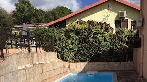Une piscine dont le fond est recouvert de carreaux de pierre, entourée d’un mur de pierre et d’une haie. On y trouve deux chaises longues, une rouge et une bleue, près de la piscine.