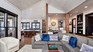 A spacious living area with a grey sofa, a white armchair, and a coffee table. A kitchen is visible in the background with a large clock on the wall.