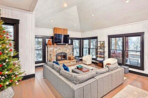 A modern living room with a stone fireplace, large windows, and a Christmas tree. - Luxury Ski In / Ski Out | Hot Tub | Sleeps 10 (Whitefish)