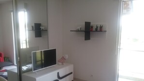 A modern living room with a flat-screen TV on a white cabinet, a glass door leading to a balcony, and a small shelf with decorative items.
