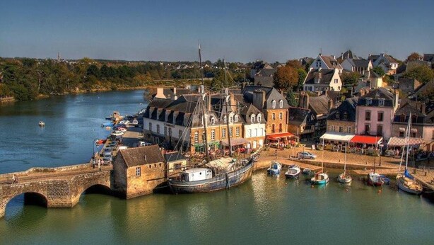 A riverside town with historic buildings, a bridge, and boats docked along the water.