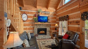 A cozy log cabin interior with a fireplace, leather sofas, a TV, and a 'Welcome' sign. - Bare Snugglin' 1 bedroom, sleeps 4! (Sevierville)