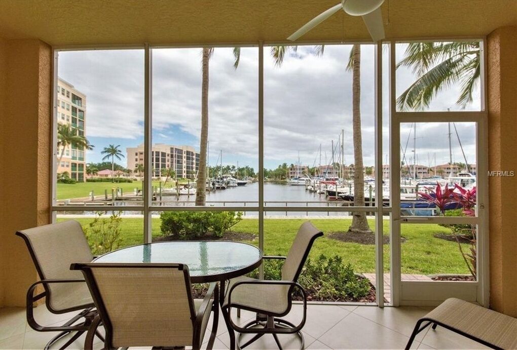 A room with a glass table and chairs, a view of a marina with boats, and a palm tree outside.