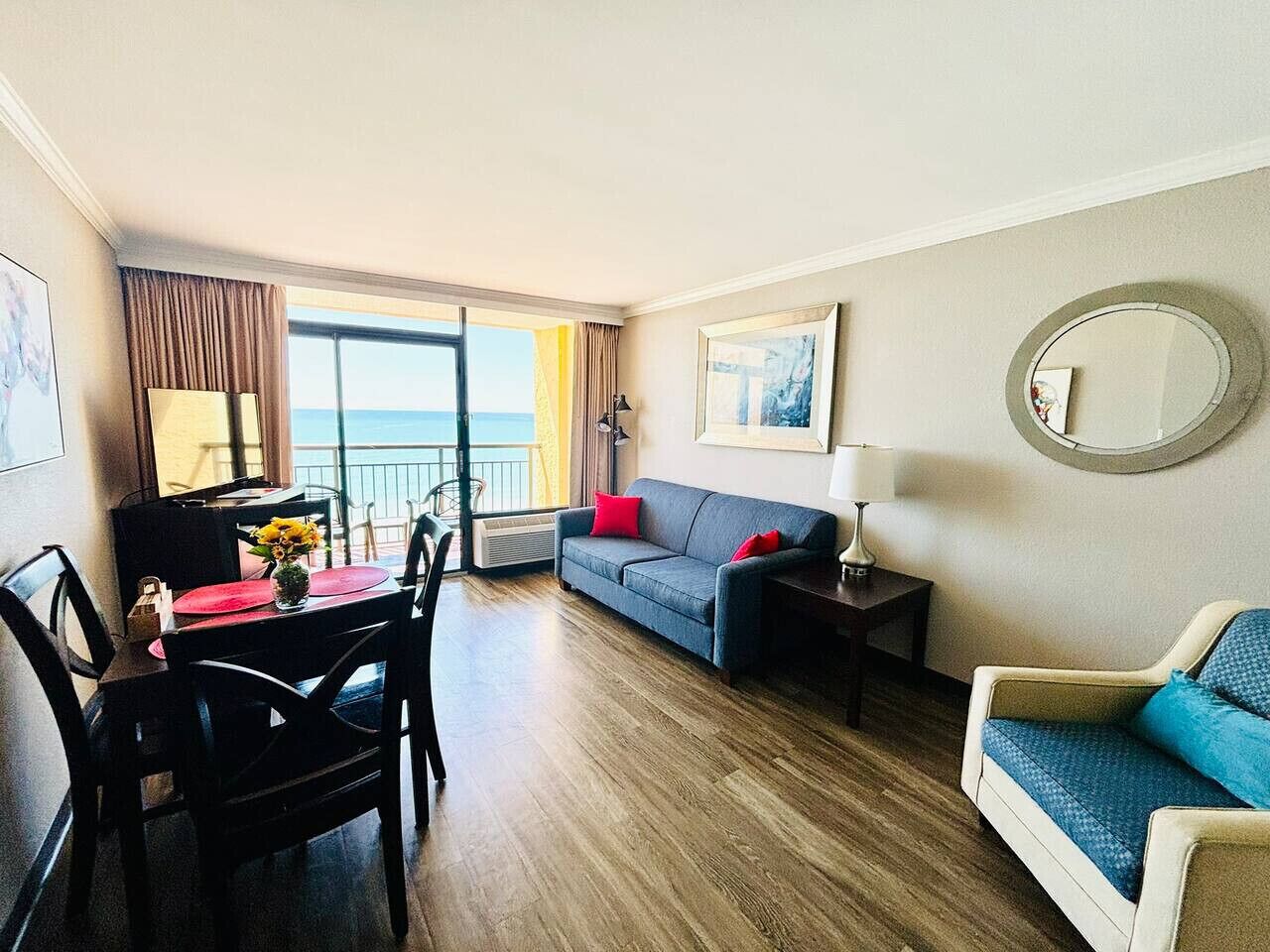 A hotel room with a dining area, a sofa, a chair, a small table with a vase of flowers, and a view of the sea through large windows.