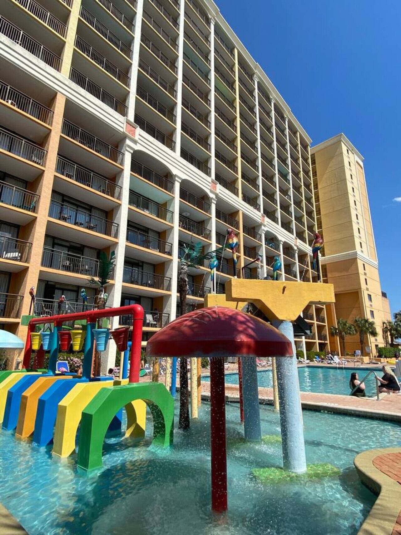Ein Hotel mit mehreren Balkonen, einem Poolbereich mit Wasserspielgeräten und wolkenlosem, blauem Himmel.