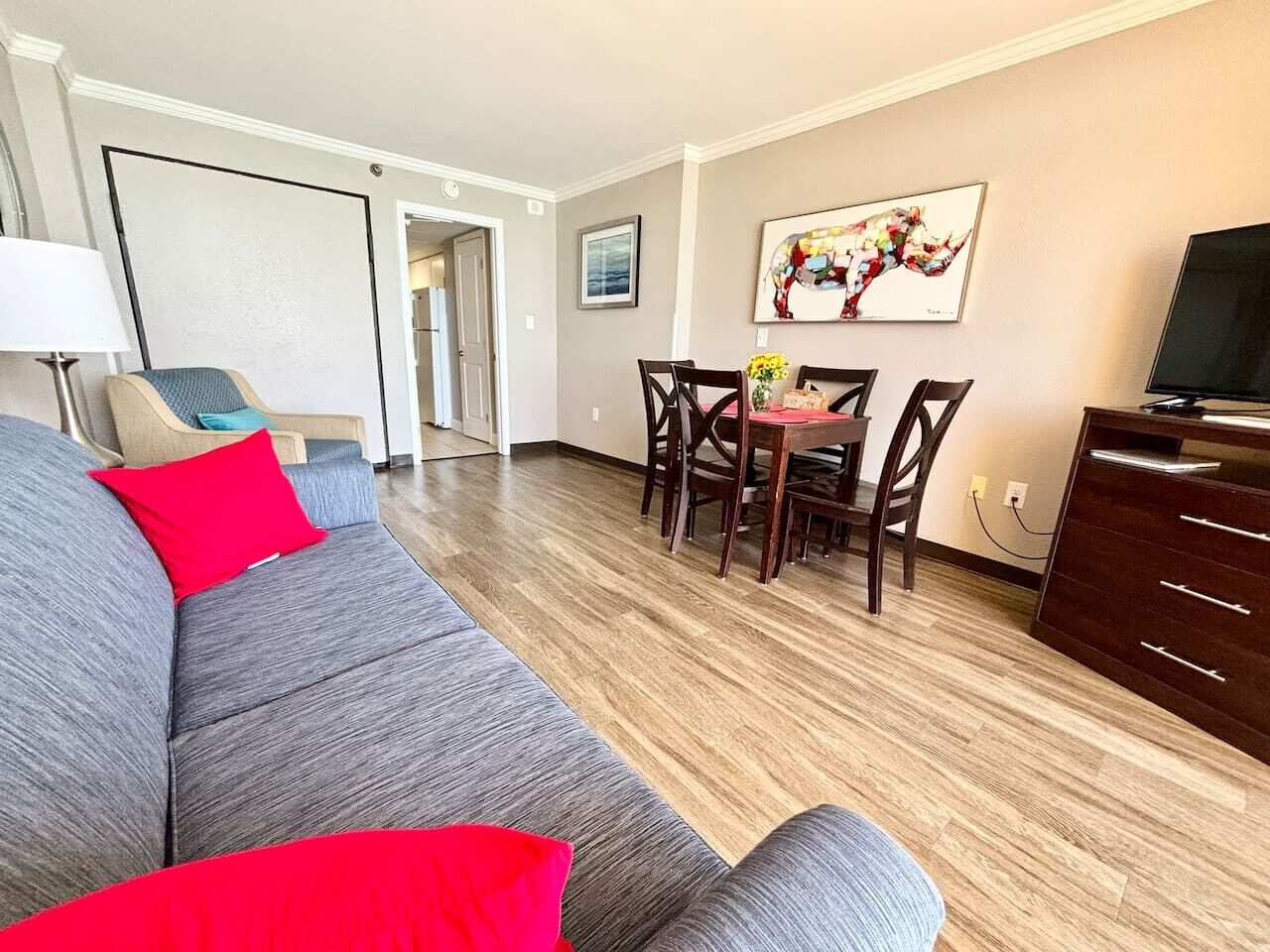 Una sala de estar moderna con un sofá gris, cojines rojos, una mesa de comedor con sillas, un mueble para televisor y un cuadro en la pared.