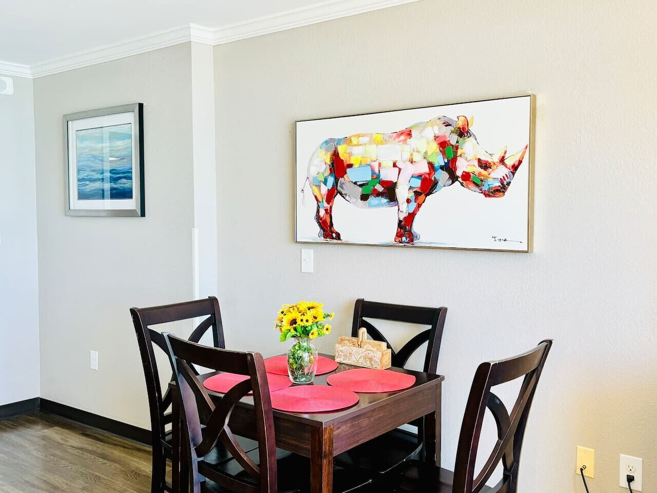 A dining area with a wooden table, four chairs, a vase with yellow flowers, and a colorful abstract painting on the wall.