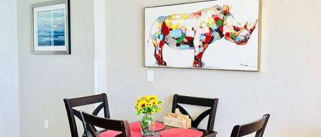A dining area with a wooden table, four chairs, a vase with yellow flowers, and a colorful abstract painting on the wall.