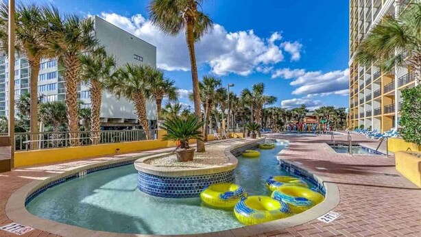 A pool area with a central fountain, surrounded by palm trees and lounge chairs. - Azure Bliss. Oceanfront Condo. Sleeps 6. (Myrtle Beach)