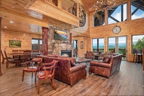 Amplia sala de estar con paredes de madera, chimenea de piedra, mobiliario de cuero y ventanas grandes con vistas al exterior.