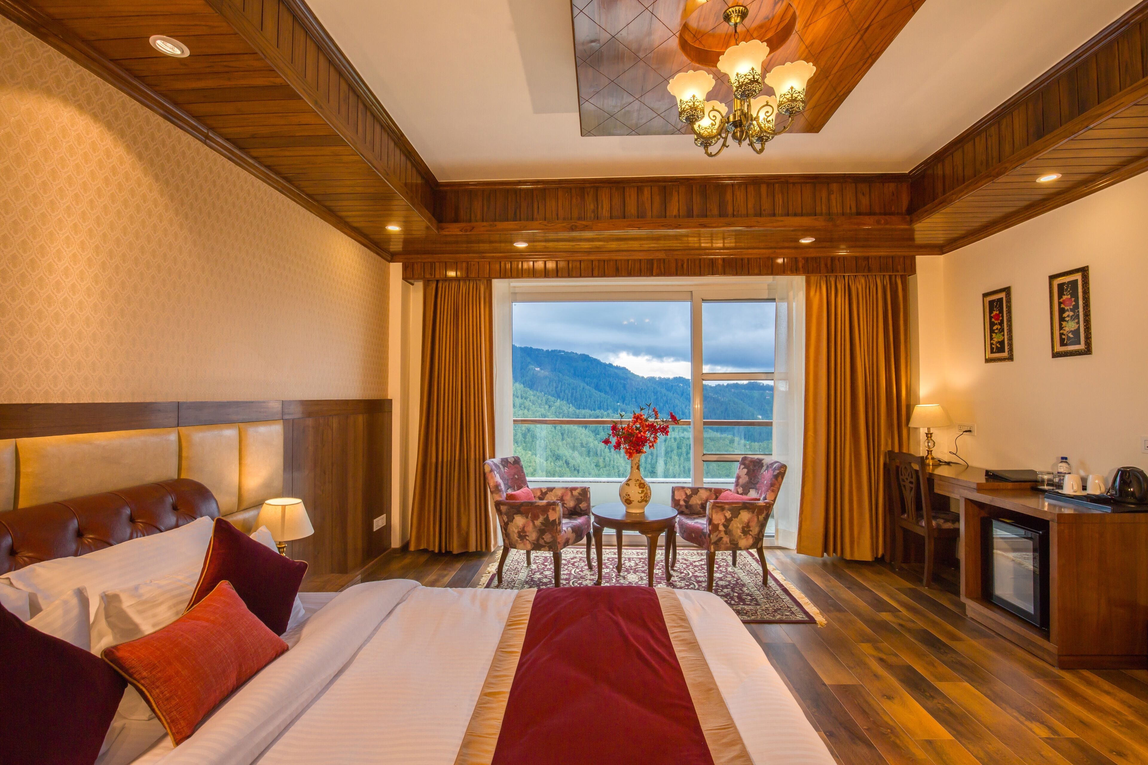 Habitación de hotel con una cama grande, un escritorio, una silla, una mesita con un florero, y vistas a las montañas desde la ventana.