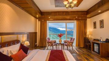 Une chambre d’hôtel avec un grand lit, un bureau, une chaise, une petite table ornée d’un vase de fleurs et une vue sur les montagnes par la fenêtre.