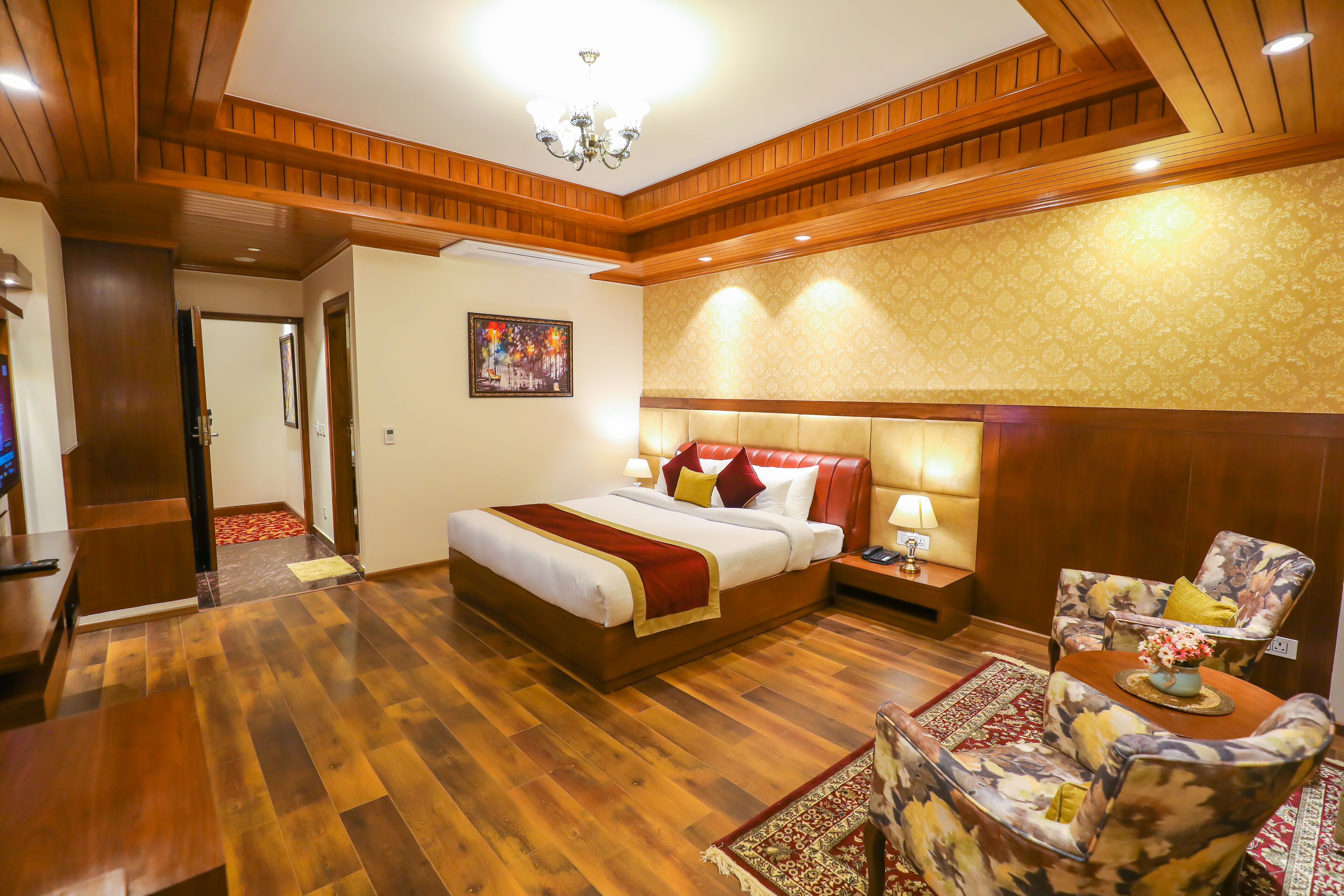 A hotel room with a wooden floor, a large bed with red and white bedding, a floral armchair, a nightstand with a lamp, and a painting on the wall.