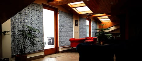 Una habitación con techo inclinado, vigas de madera, un papel tapiz estampado, una silla roja y una planta en maceta.