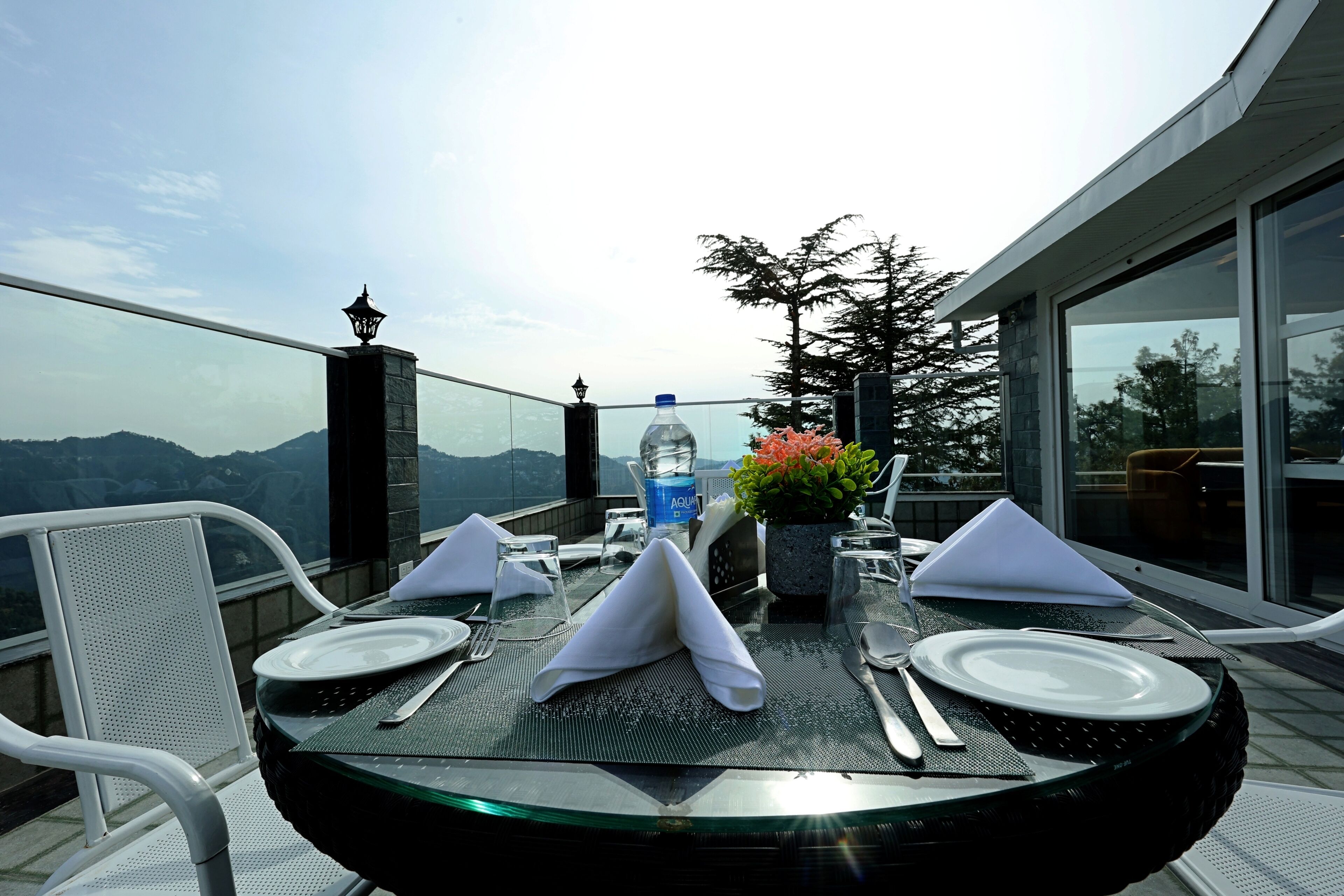 A dining table set with plates, cutlery, and a water bottle on a balcony with a mountain view.