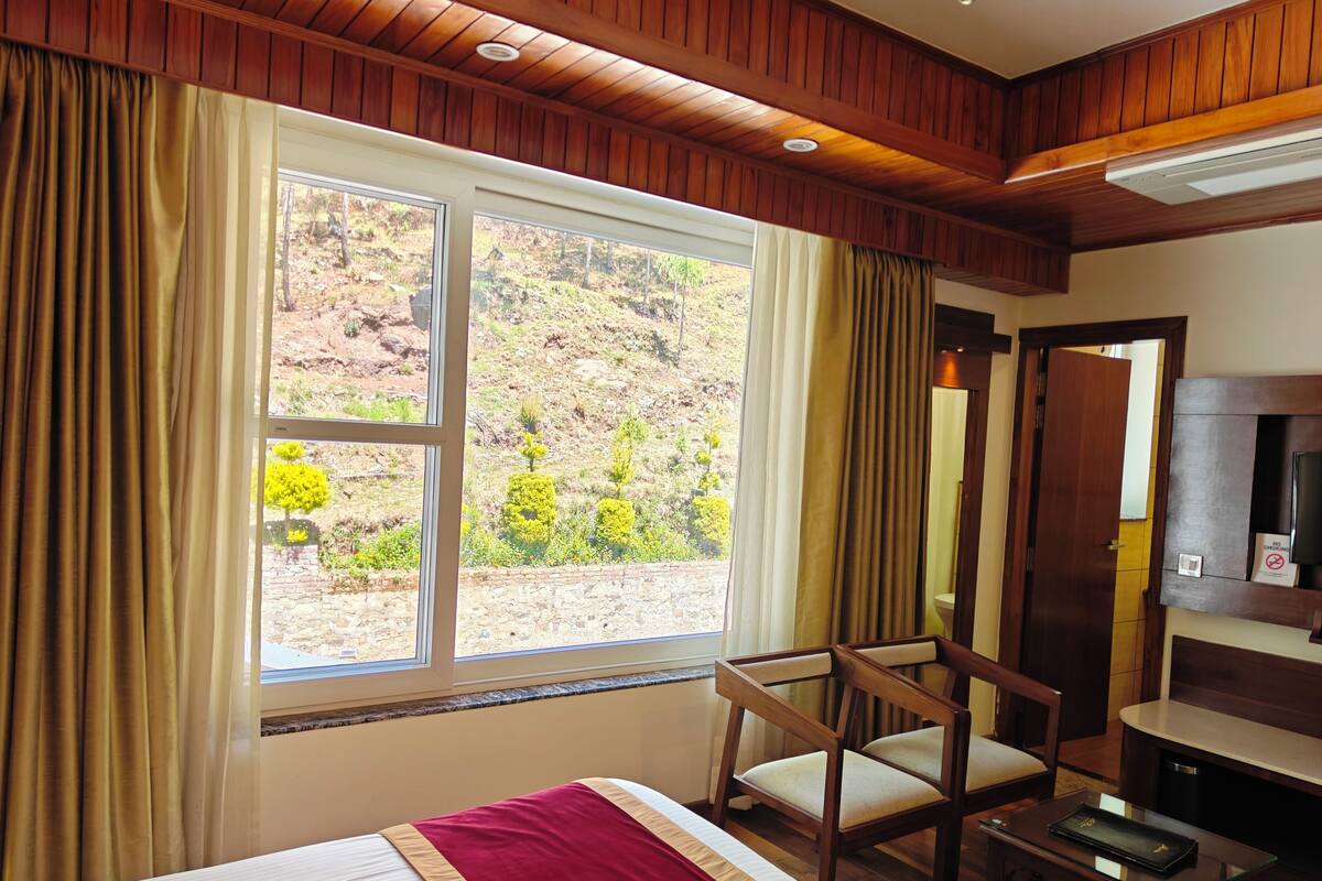 Habitación de hotel con cama, una silla de madera, un escritorio y un ventanal con cortinas.
