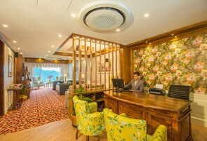 A hotel reception area with a wooden desk, two yellow floral chairs, a large floral wallpaper, and a view of a mountain through a window.