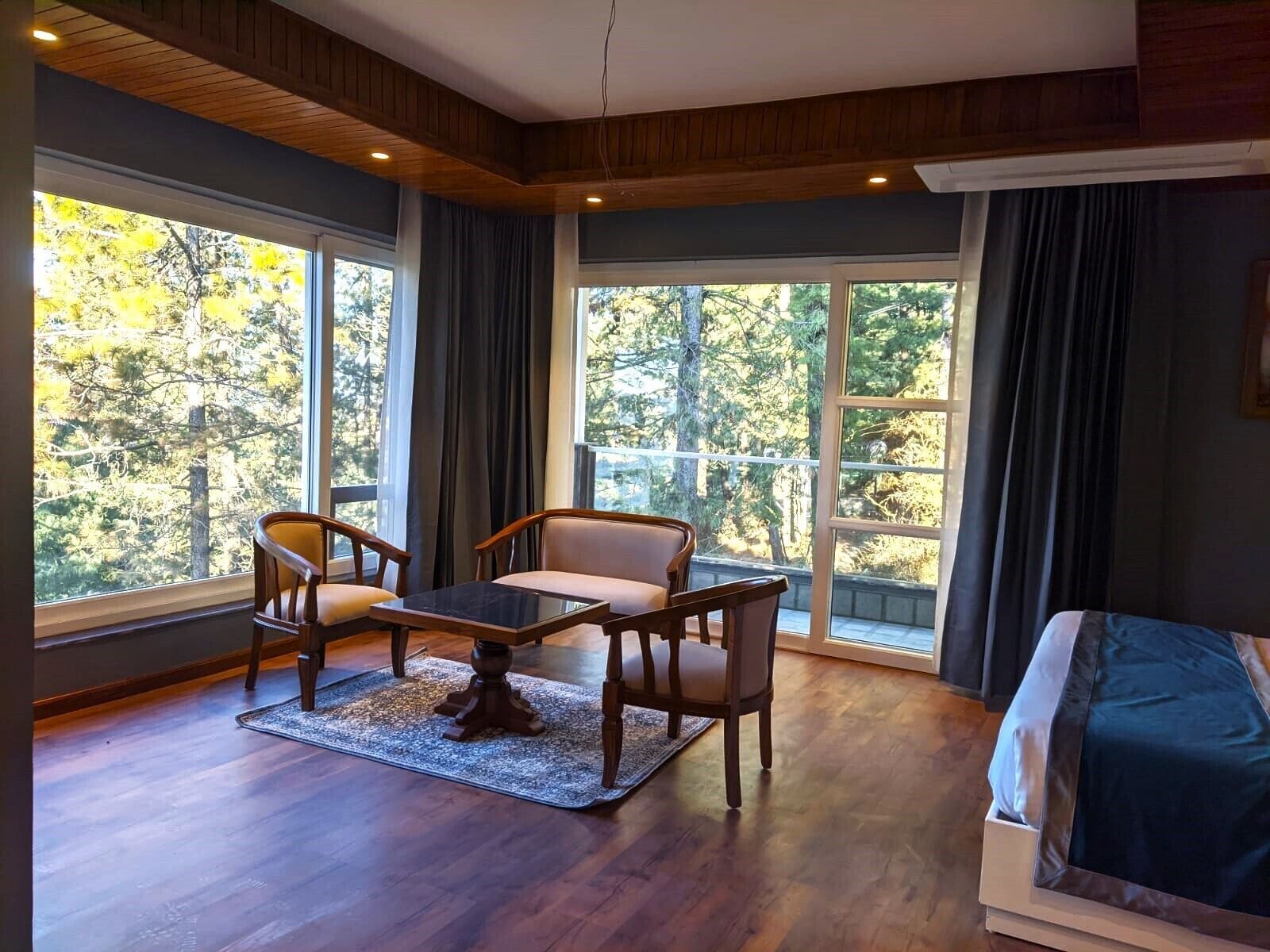Una habitación con piso de madera, un sofá, una mesa y dos sillas. Cuenta con ventanas amplias con cortinas, y se ve un paisaje de árboles desde el interior.