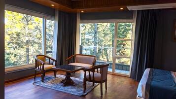 Une pièce avec un plancher en bois, un canapé, une table et deux chaises. De grandes fenêtres, ornées de rideaux, offrent une vue sur les arbres à l’extérieur.