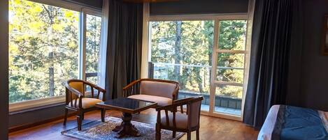 A room with a wooden floor, a sofa, a table, and two chairs. There are large windows with curtains, and a view of trees outside.