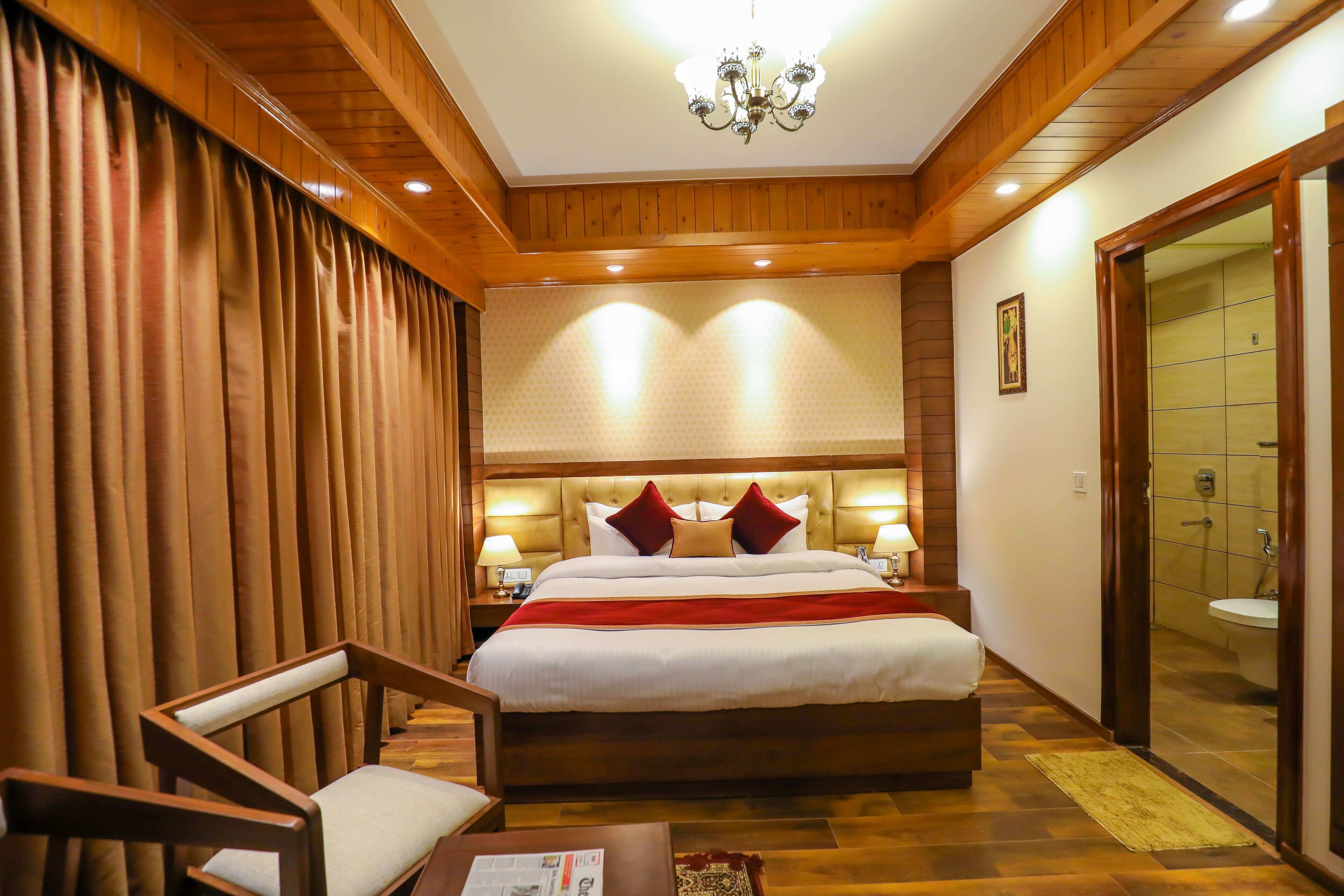 Una habitación de hotel con una cama grande, paneles de madera, una araña, una mesita de noche con lámparas y un baño visible a través de una puerta abierta.