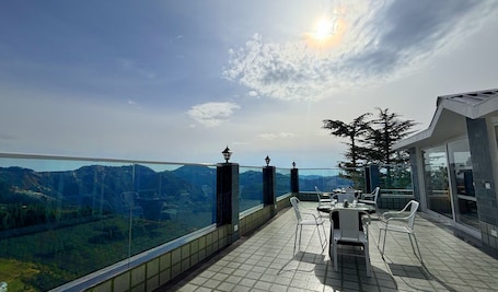 A terrace with a panoramic view of mountains, a dining table with chairs, and a glass railing.
