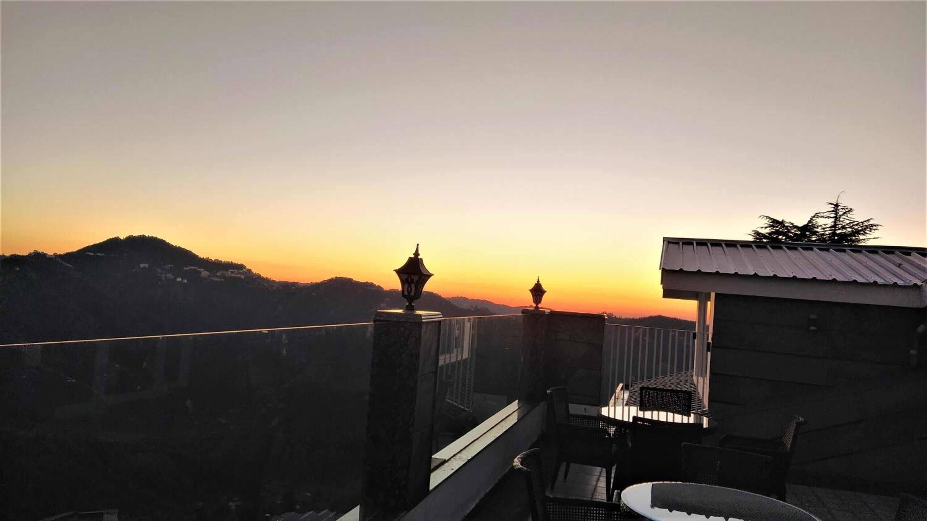 A balcony with a view of a sunset over a mountainous landscape, a small table, and a chair.