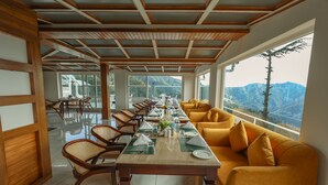 A dining area with a long table set for a meal, wooden ceiling beams, and a view of mountains through large windows. - The Retreat Mashobra, Shimla (Shimla)