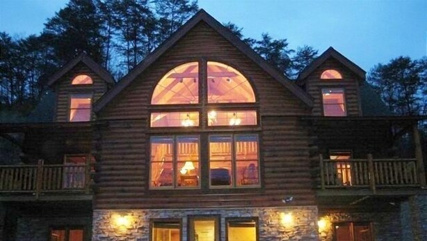 Una cabaña de troncos con base de piedra, amplias ventanas arqueadas y un balcón.