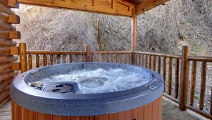 Un jacuzzi en una terraza de madera con barandilla de madera y una cabaña de troncos al fondo.