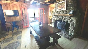 Una stanza accogliente con camino in pietra, travi a vista in legno, un ampio tavolo in legno e un televisore fissato al muro.