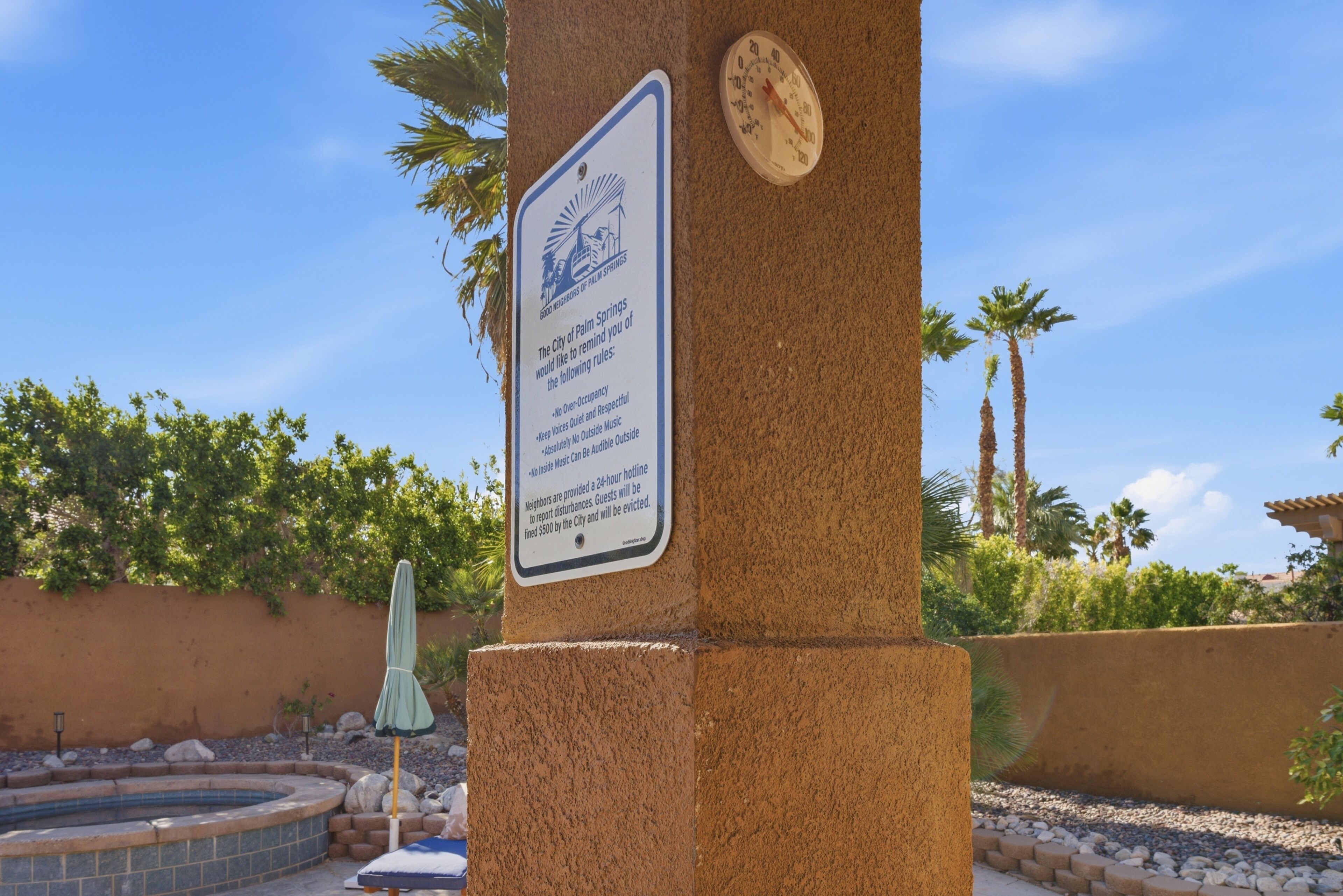 A sign with rules for Palm Springs, a thermometer, and a pool area with rocks and greenery.