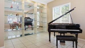 Un piano à queue dans une pièce bien éclairée, dotée d’un sol carrelé, d’étagères en verre ornées d’objets décoratifs et, au fond, d’un coin repas.